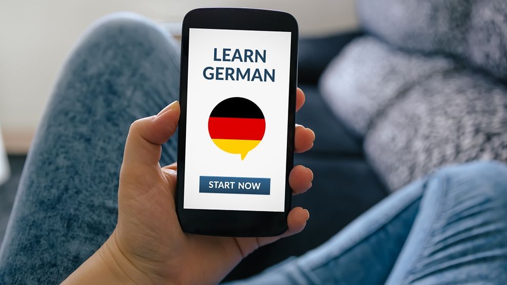  A woman sitting on a sofa and holding a smartphone with a learn german app on screen. 